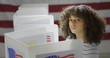 © vesperstock - MS Young Hispanic woman in polling station, voting in a booth with US flag in background. Serious expression from high viewpoint