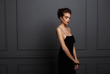 Profile Portrait Of A Sensual Brunette Woman In Sexy Black Dress With Bare Shoulders And Beautiful Earrings Near Grey Dark Neoclassical Wall.