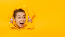 Horizontal Banner Of Young Female Tearing Paper And Peeking Out Hole, Curious About Commercial Offer On Copy Space On Right, Isolated On Yellow Background