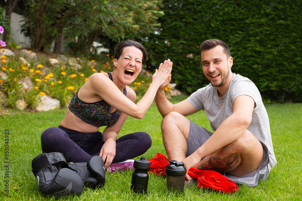 Joyful couple of fighters enjoying break during outdoor workout, sitting on grass, giving high five. Exercising outdoors and trainer job concept. - obrazy, fototapety, plakaty 