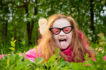 Playful funny little girl with long hair and in pink glasses having rest and fun on green grass in a summer park