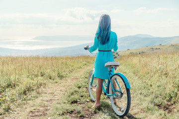 Wall Mural - Girl with blue hair standing with bicycle on country road along summer meadows.