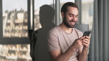 Canvas Print - A smiling pleased young man is using his smartphone sitting at home in the living room