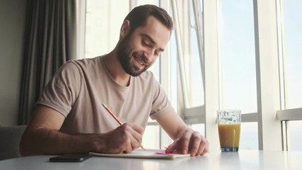 Poster - A pleased young man is writing something by hand sitting at the table at home 