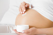Girl sitting in bed. Young woman hands holding opened white cream jar and applying naked big belly. Care about perfect, soft and smooth skin in pregnancy time. Close up. Side view.