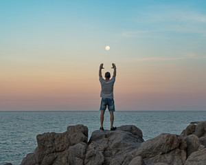 Wall Mural - Young man in casual clothes stands on the rocks by the sea at sunset and the full moon between his arms raised 