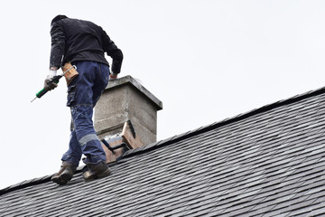 Roofer construction worker repairing chimney on grey slate shingles roof of domestic house, sky background with copy space.
