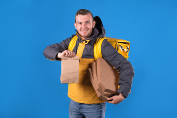 Portrait of unshaven guy courier smiling in yellow uniform holding pizza boxes and food in paper bag and thermobag blue background. Concept of fast, delicious and high-quality food delivery.