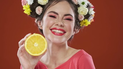 Poster - A cheerful and emotional brunette woman with an amazing floral hairstyle is posing with an orange isolated over a red background in studio