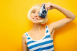 © moisevich - Portrait of a smilling girl with a camera in hand on a yellow background. Isolated studio. blonde wig