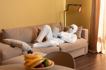 KYIV, UKRAINE - APRIL 24, 2020: Selective focus of man in hazmat suit and medical mask using laptop near joystick and remote controller on couch