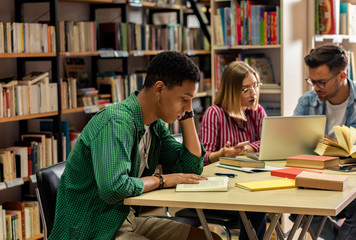 Wall Mural - Portrait of young african american student sitting in library studying, with his friends in background.