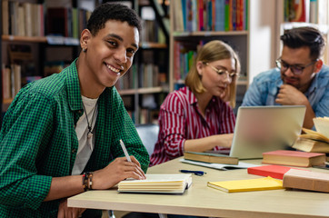 Wall Mural - Portrait of young african american student sitting in library looking at camera, with his friends in background.