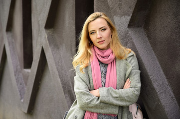 Portrait of a caucasian blonde woman in a coat. A model stands outdoors against the background of a wall of a building in the city.