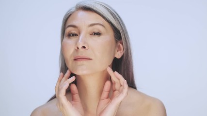 Wall Mural - A good-looking old mature half-naked woman with long gray hair is looking to the camera like in a mirror and touching her skin isolated over white background in studio