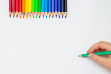 Wall Mural - Top view of left hand of child holding pencil and drawing on white paper with the set of colour pencils. Copy space.