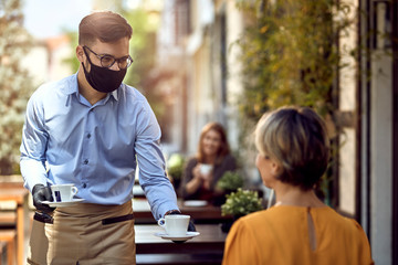 Happy waiter wearing protective face mask while serving coffee to his guest.