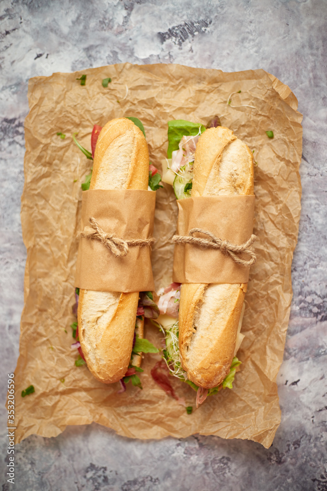 Sandwiches with ham, fresh vegetables and herbs served on brown baking paper over concrete backdrop - obrazy, fototapety, plakaty 