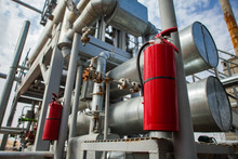 Close-up Of Red Fire Extinguisher On Oil Refinery Plant. Grey Pipes And Pipe-lines And Blue Sky With Clouds On Background. CNPC Company.