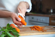 Hands of a young woman in an apron and on the background of the kitchen, peeling vegetables using a food peeler. Cleans the peel from carrots. Cooking fresh carrots before serving.