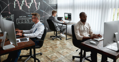 Poster - A world for learners. Three traders sitting by desks in front of computer monitors while working in the office. Blackboard full of charts and data analyses in background