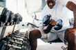 © twinsterphoto - Young African American man sitting and lifting a dumbbell close to the rack at gym. Male weight training person doing a biceps curl in fitness center