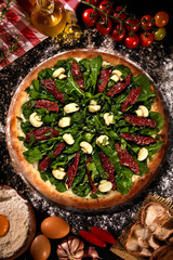 Wall Mural - Tasty arugula and dried tomato pizza and cooking ingredients tomatoes basil on wood background. Top view of arugula pizza