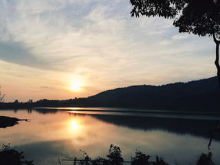 Serene sunset over a calm lake with golden sunlight reflecting on the water, silhouetted mountains, and a peaceful natural landscape at Bang Wat Dam, Phuket, Thailand.