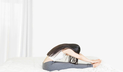 young asian woman wearing white undershirt and gray pants,exercising yoga while sitting on a white bed , stretch oneself after waking up at home Health care concep,selective focus