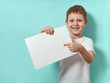 © fascinadora - Four-year-old boy smiles and shows finger on blank white sheet. Happy child on blue background with copy space for message, mock up