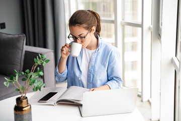 Canvas Print - Image of woman reading book and drinking tea while working with lapto