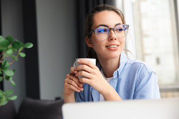 Wall Mural - Image of woman drinking tea and working with laptop while sitting at table