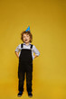 Stylish little boy in overalls and birthday cap posing at the yellow background, isolated. Kids fashion