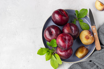 Wall Mural - Whole purple plums and slices with leaves and knife on ceramic plate, light grey concrete background. Top view.