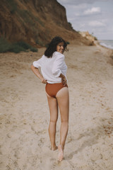 Boho girl in white shirt running on sunny beach, back view. Carefree stylish woman in swimsuit and shirt relaxing on seashore. Summer vacation. Lifestyle authentic image