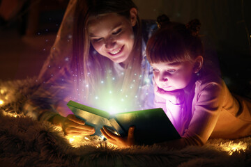 Wall Mural - Beautiful young woman and her little daughter reading book at home in evening