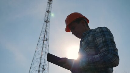 A silhouette of a telecommunications network engineer in a hard hat uses a tablet at work.