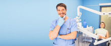 Young Smiling Handsome Male Doctor In Blue Medical Uniform, Disposable Medical Facial Mask With Equipment In Dentistry Office And Happy Patient In The Dental Chair. Stomatology Concept. Copy Space