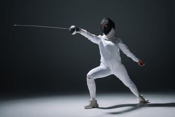 Swordswoman in fencing mask and suit holding rapier on white surface on black background