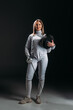 Beautiful fencer looking at camera while holding rapier and fencing mask on grey surface on black background