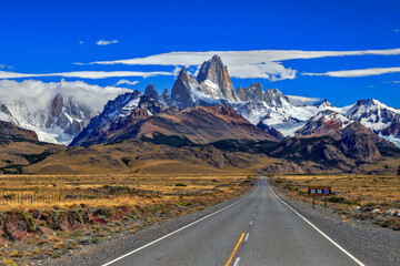 Fitz Roy and Torre mountains with route and cars at foreground, during a sunny day with clouds.