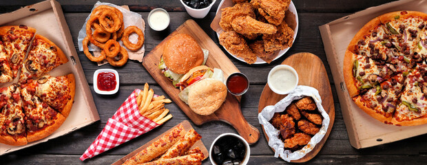 Wall Mural - Table scene of assorted take out or delivery foods. Hamburgers, pizza, fried chicken and sides. Top down view on a dark wood banner background.
