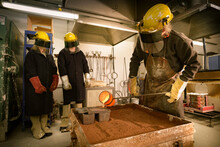 Three People Wearing Overalls And Protective Equipment Watching One Pouring Molten Metal Into A Mold.