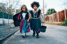 Two Girls Dressed For Halloween Walking Down A Street Holding Hands.