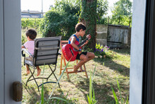 Two Boys Sitting On Chairs In The Garden, One Playing A Guitar.