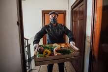 Man Wearing Flat Cap And Face Mask Delivering Vegetable Box In An Apartment Building During Corona Virus Crisis.