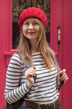 Outdoor Portrait Of Young Beautiful Woman In Striped Shirt And Red Beret. A Lady Is Dressed In The French Style. In The Background Is A Red Door.