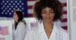 © vesperstock - CU Young mixed-race woman in white lab coat with 'I Voted' sticker, in front of voting booths in US election. Other scientists/doctors of various demographics, in white lab coats, vote in background.