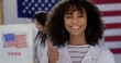 © vesperstock - CU Young Hispanic woman in white lab coat with 'I Voted' sticker, smiles and gives thumbs up. Other scientists of various demographics, in white lab coats, vote in background.