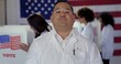© vesperstock - CU Hispanic man in white lab coat with 'I Voted' sticker, standing at polling station. Other scientists/doctors of various demographics, in white lab coats, vote in background.